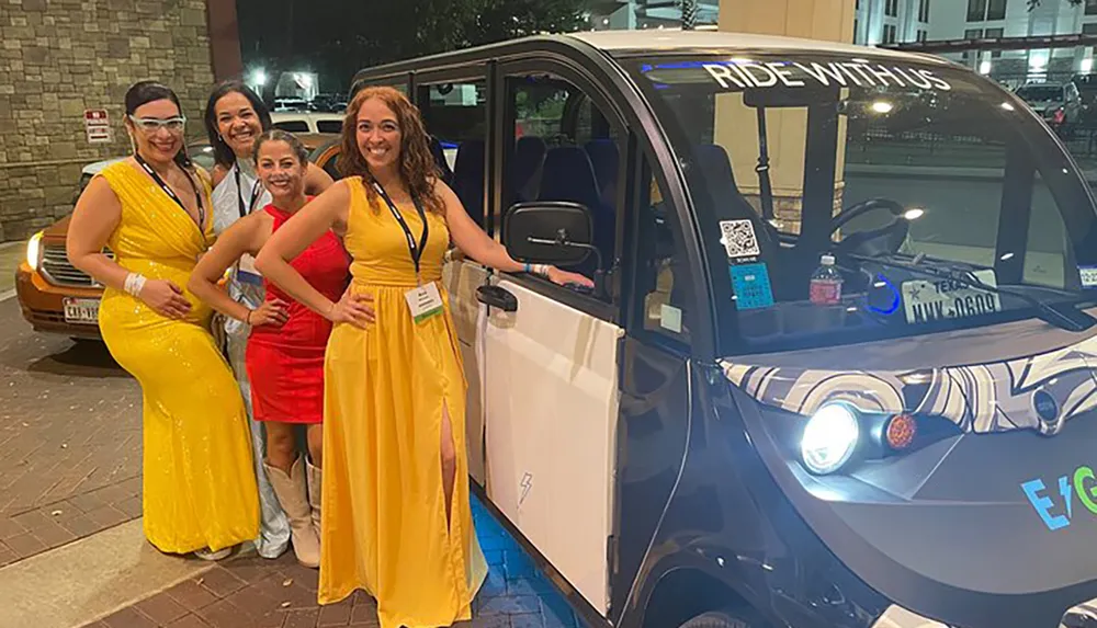 Four smiling women in semi-formal attire are posing next to a small modern passenger shuttle vehicle at night