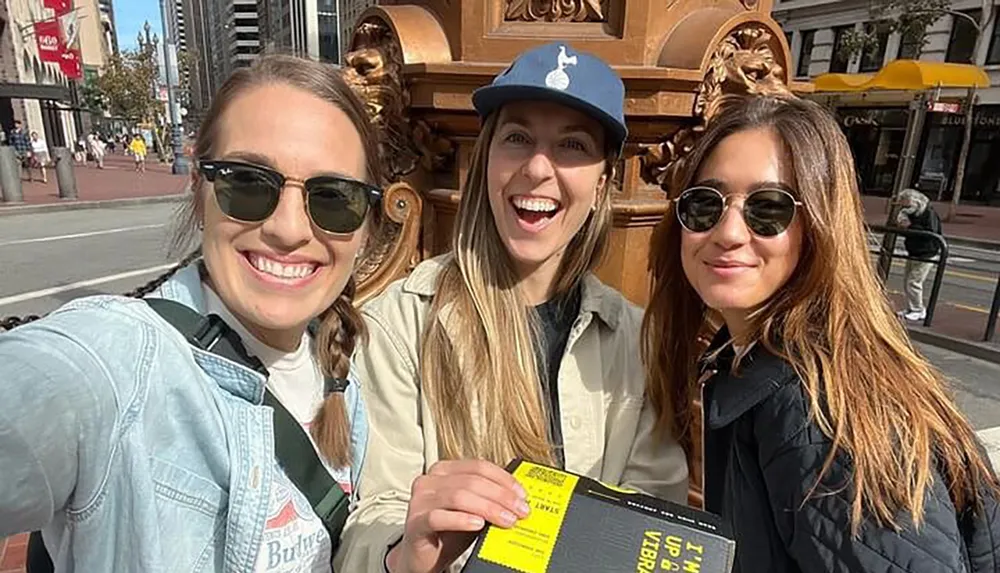 Three smiling women are taking a selfie on a sunny day in an urban setting with one of them holding a book titled Im Glad My Mom Died