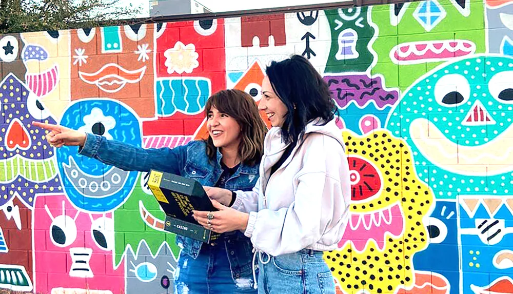 Two people are smiling and one is pointing at something off-camera standing in front of a vibrant multicolored mural with playful abstract designs