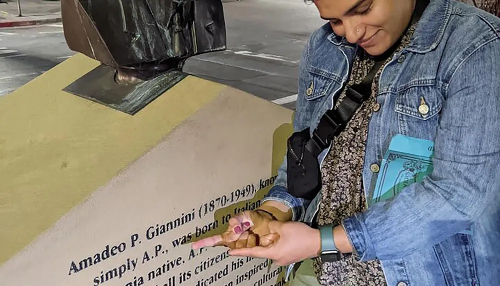 A person is reading a plaque about Amadeo P Giannini while holding a camera standing in front of a pedestal with a bust on it