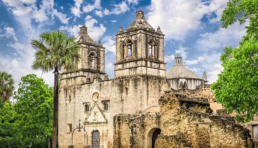 The image shows an old weathered mission with twin bell towers and a dome surrounded by green foliage under a partly cloudy sky