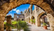 This image shows a view of an old stone mission with arches, a dome, and a bell tower, framed by a weathered stone archway under a clear blue sky.