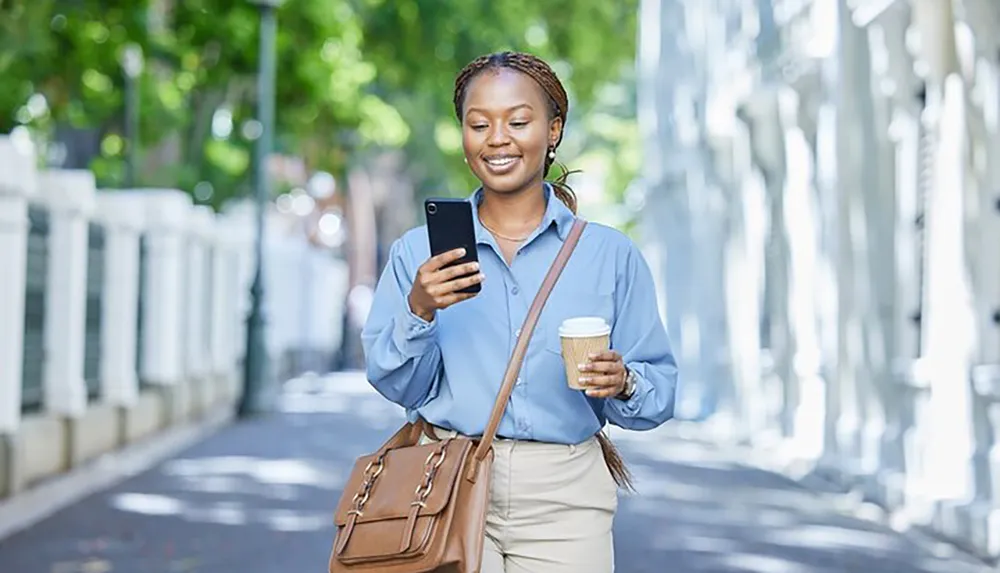 A smiling person is walking on a tree-lined street looking at a smartphone in one hand with a coffee cup in the other