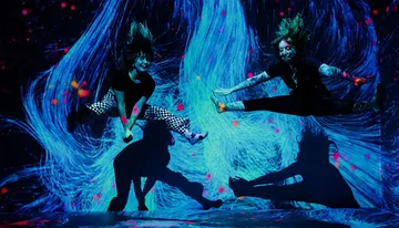 Two dancers are captured in mid-air against a vibrant blue and pink neon-lit background, creating a dynamic sense of movement and energy.