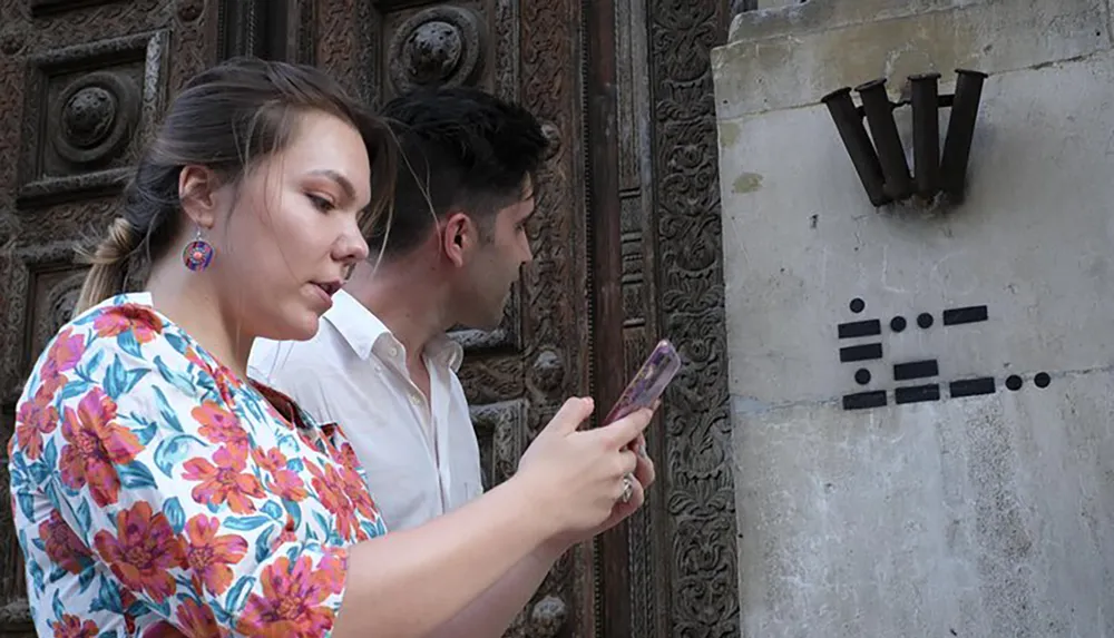 A woman is examining her phone while standing next to a man with both of them seemingly ignoring or unaware of the artistic arrangement of metal objects on the wall beside them