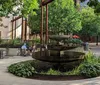 A person in a yellow dress is walking past a tiered fountain in a peaceful urban park setting with seated individuals and lush greenery