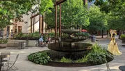 A person in a yellow dress is walking past a tiered fountain in a peaceful urban park setting with seated individuals and lush greenery.