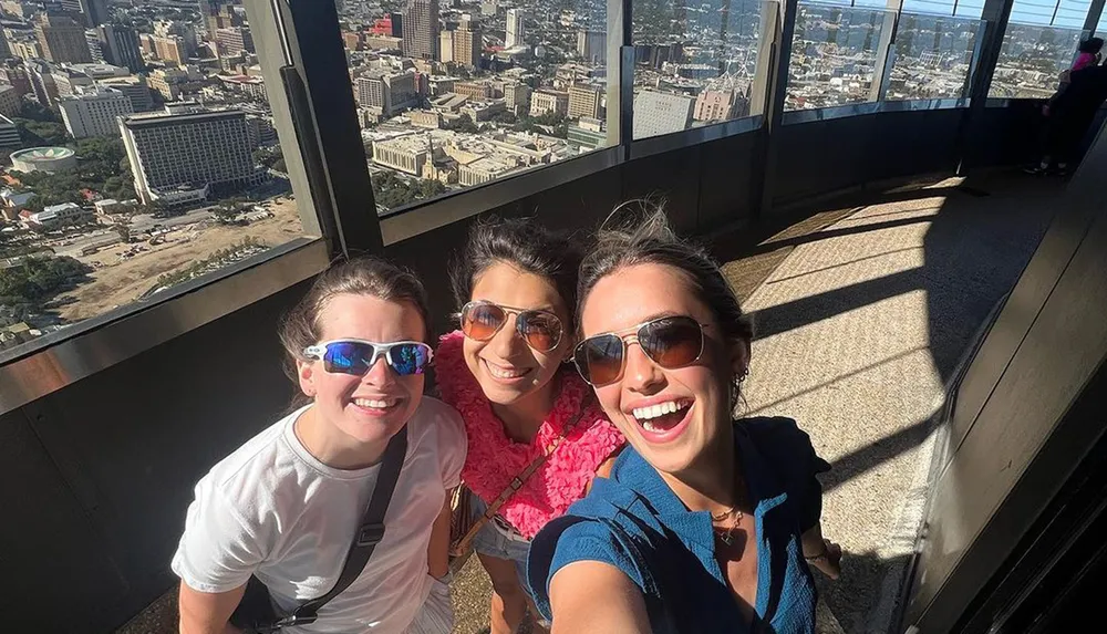 Three people are smiling for a selfie with a cityscape visible from a high vantage point in the background