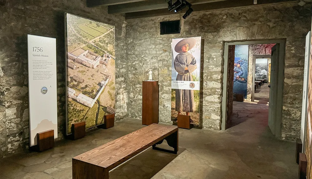 The image shows an exhibition room with stone walls featuring display panels a wooden bench and educational material related to a Spanish mission from 1756