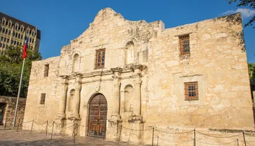 The image shows the historic Alamo mission in San Antonio, Texas, with its distinctive facade under a bright blue sky.