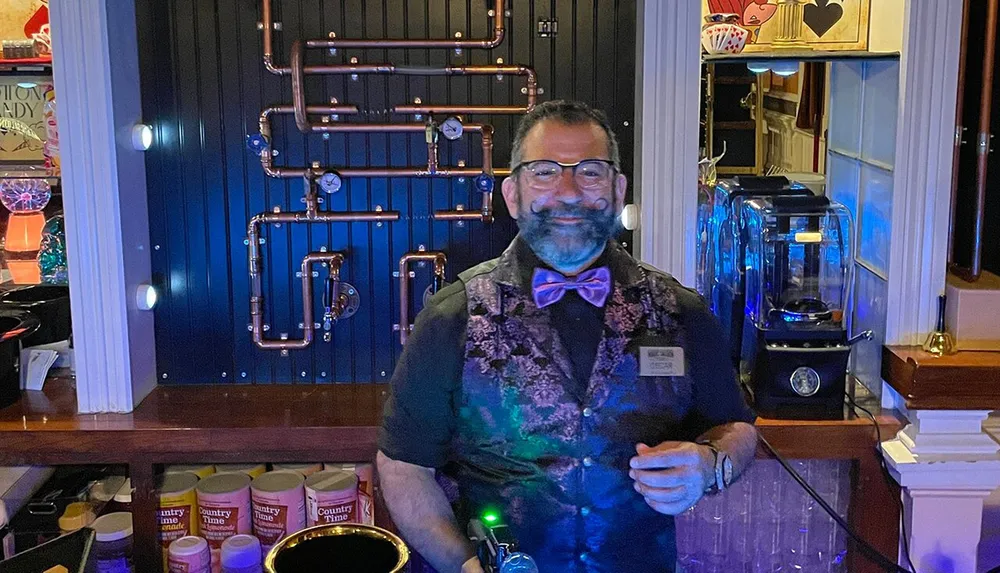 A man wearing a bow tie and vest stands behind a bar with industrial pipe decor on the wall behind him