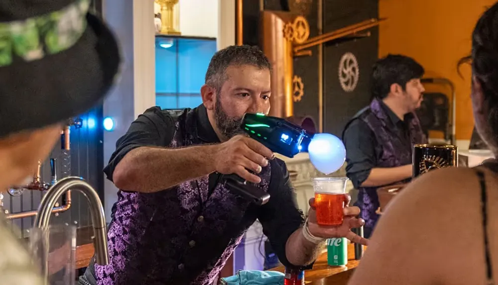 A man is using a futuristic device to prepare a drink while wearing a vest behind a bar with other people nearby
