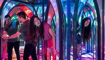 Several people navigate a colorful and reflective mirror maze, with vibrant lights creating a kaleidoscopic effect.