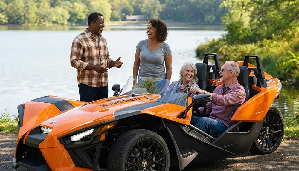 Four people are smiling and talking by a lake with two of them seated in an orange three-wheeled vehicle