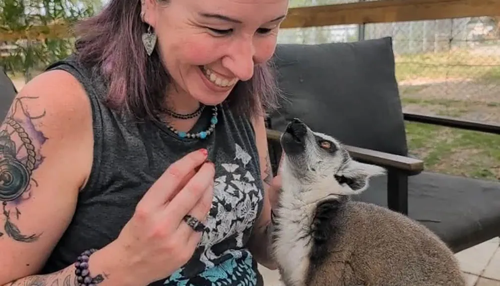 A woman with tattoos and jewelry is smiling at a lemur looking up at her
