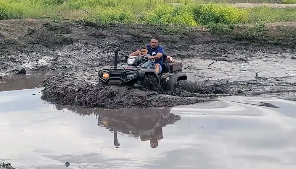 Two people are riding an ATV through a muddy patch of land creating splashes and tracks in the mud