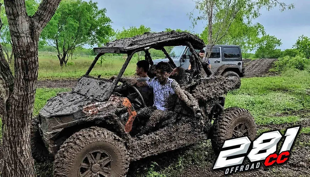A muddy off-road vehicle carrying two people drives through a grassy area with a jeep in the background