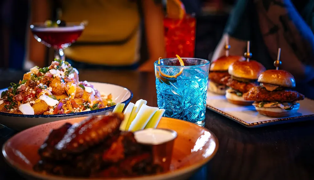 The image shows a variety of appetizing dishes and colorful cocktails on a dark wooden table suggesting a vibrant dining experience