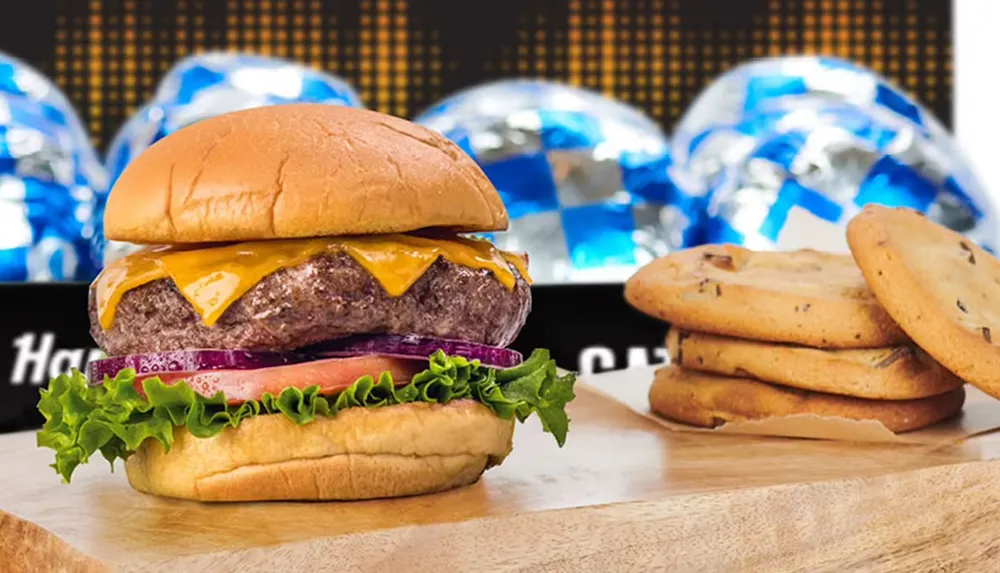 A cheeseburger with lettuce tomato and onion is prominently featured alongside a stack of chocolate chip cookies and foil-wrapped items in the background