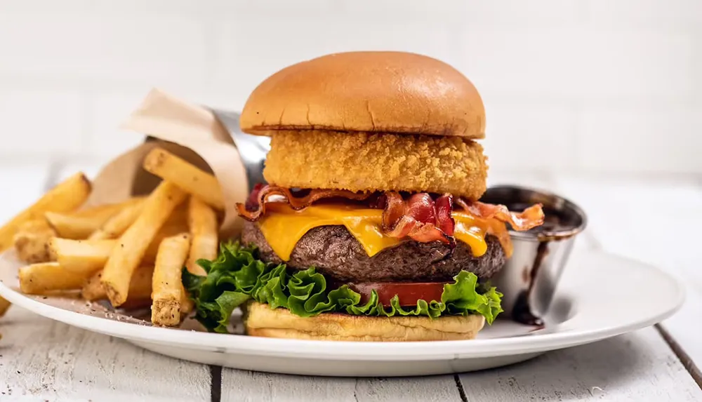 The image shows a hearty cheeseburger with a beef patty lettuce bacon cheese and a crispy onion ring accompanied by French fries and barbecue sauce on a white plate against a light background