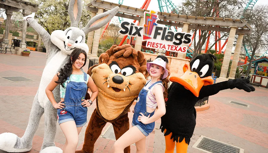 Two people are posing with costumed characters Bugs Bunny, Taz, and Daffy Duck at the Six Flags Fiesta Texas theme park.
