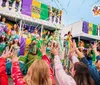 A festive Mardi Gras parade features colorful performers on a float with a lively crowd reaching up towards them