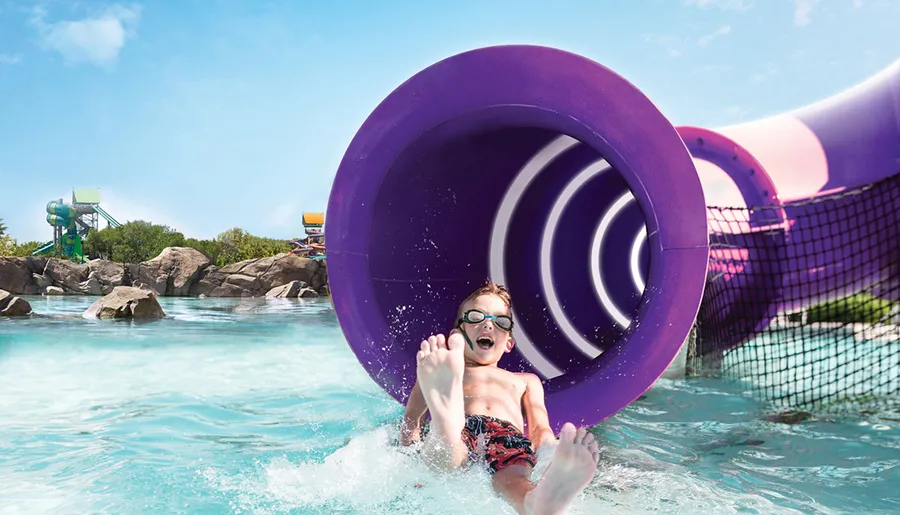 A child with goggles is sliding out of a purple water slide into a splash pool with an expression of excitement.