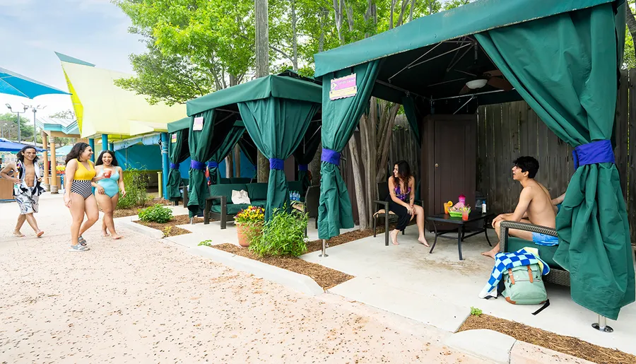 People are enjoying a day at a water park, with some relaxing in cabanas and others walking by in swimwear.