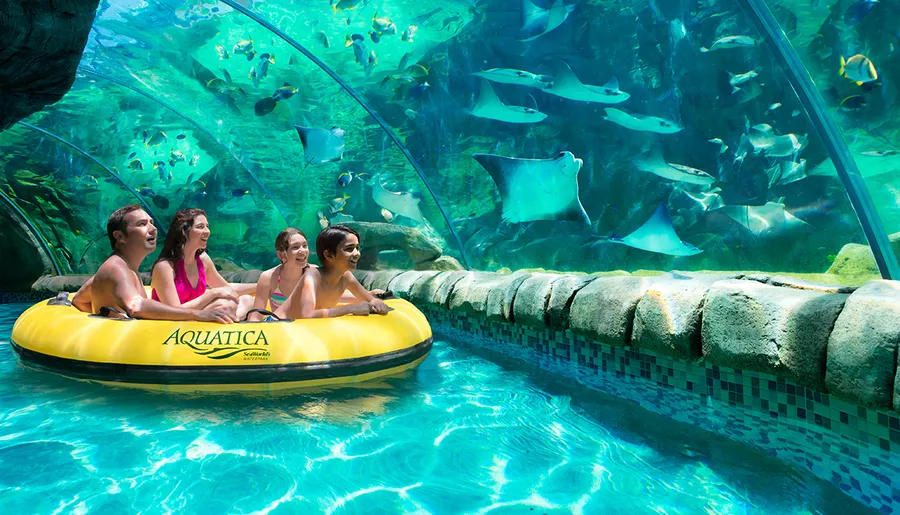 A family enjoys a ride on a yellow inflatable raft inside a water tunnel while observing aquatic life swimming above them.