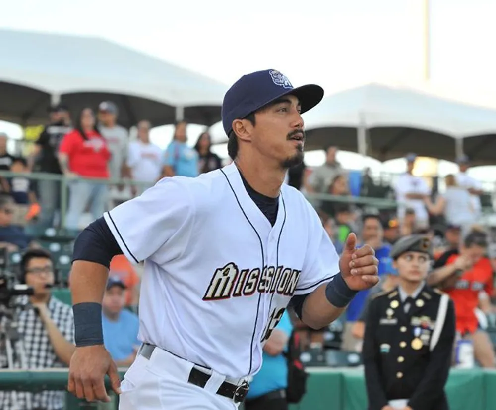 A baseball player is gesturing with his hand while looking out onto the field wearing a jersey with the word MISSIONS printed on it with spectators and a uniformed guard in the background