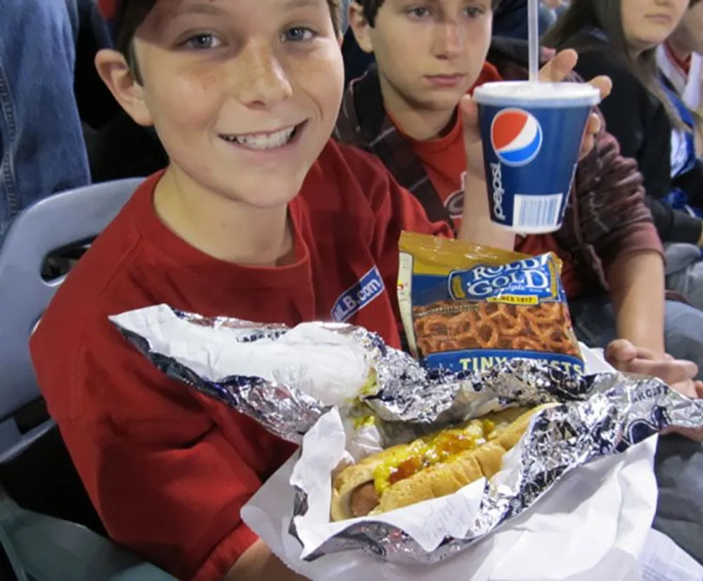 A smiling young person is holding up a hot dog topped with mustard accompanied by a bag of pretzels and a cup of Pepsi likely enjoying a sporting event