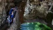 Visitors are exploring a naturally illuminated cave with a clear pool of water and intriguing rock formations.