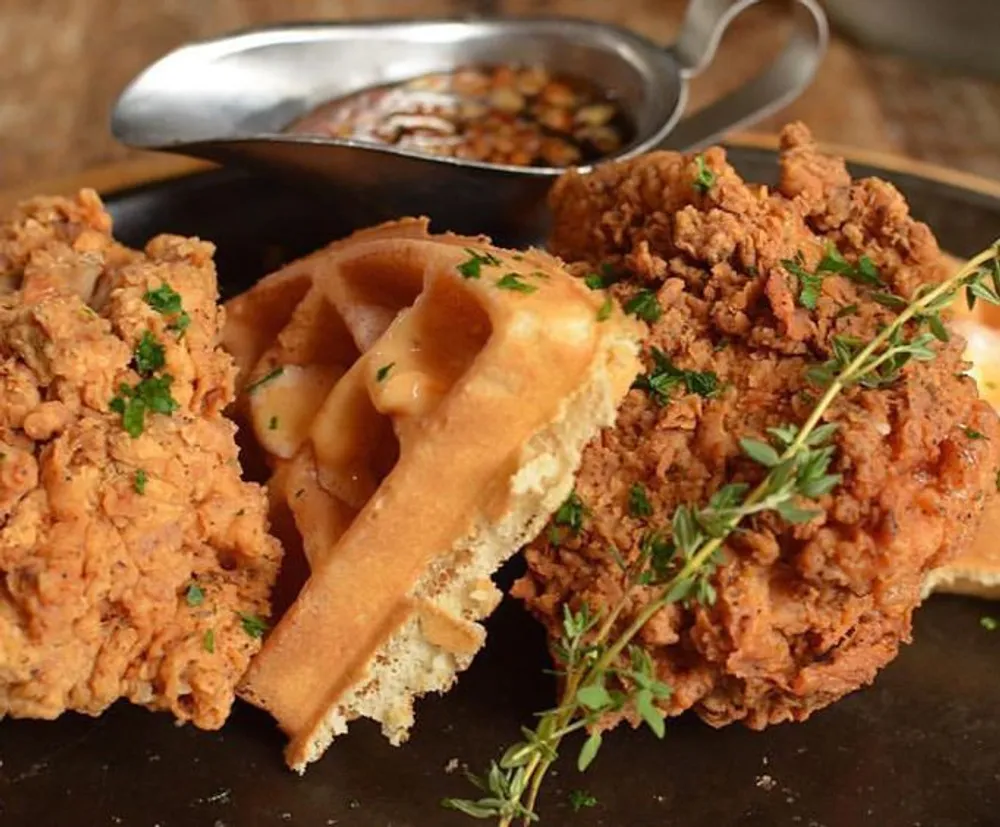 The image shows a plate of crispy fried chicken paired with a waffle drizzled with sauce and garnished with herbs indicating a savory and sweet dish combination