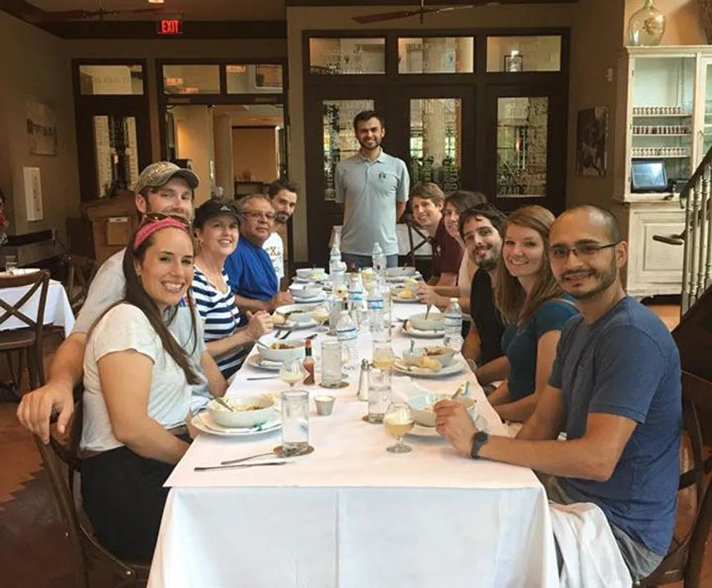 A group of smiling people is gathered around a dining table in what looks to be a casual restaurant setting suggesting a social or familial gathering