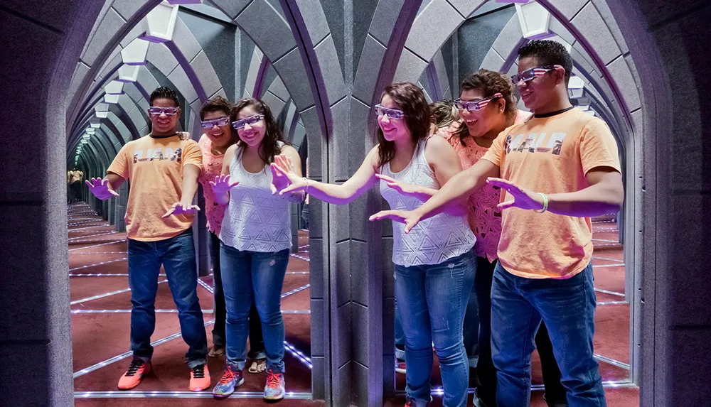 A group of people are wearing 3D glasses and posing with their hands out in a futuristic corridor with mirrored reflections extending the space infinitely