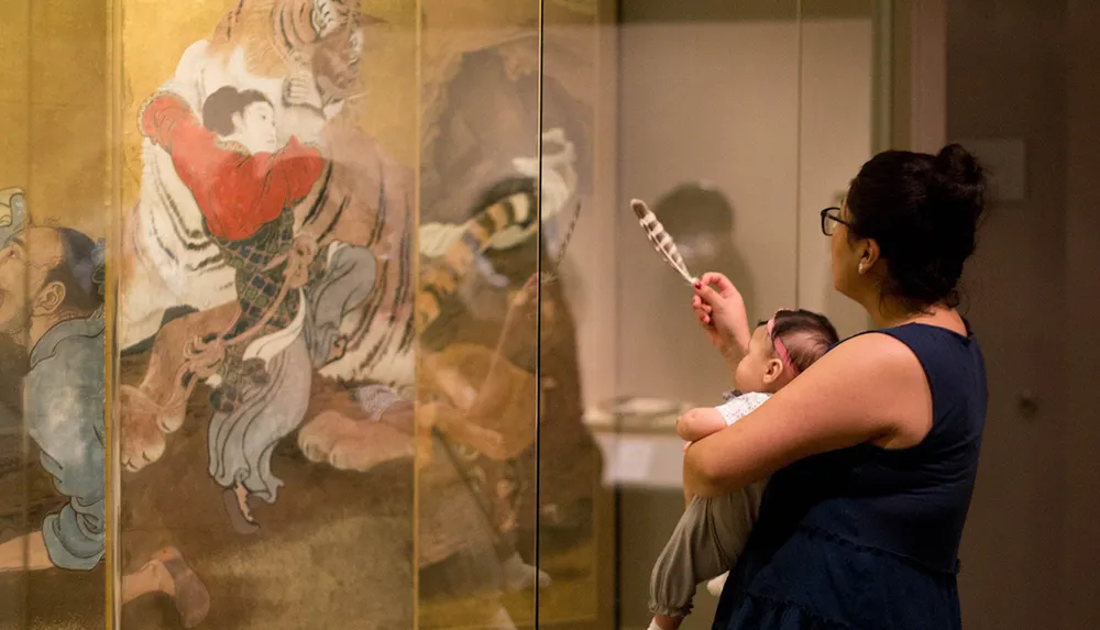 A woman is holding a baby and pointing at an Asian-style painting or artwork of a man wrestling a tiger as they look at it displayed behind glass in a museum