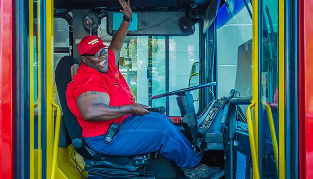 A smiling bus driver in a red shirt and cap is seated at the wheel of a bus waving to the camera with a friendly gesture