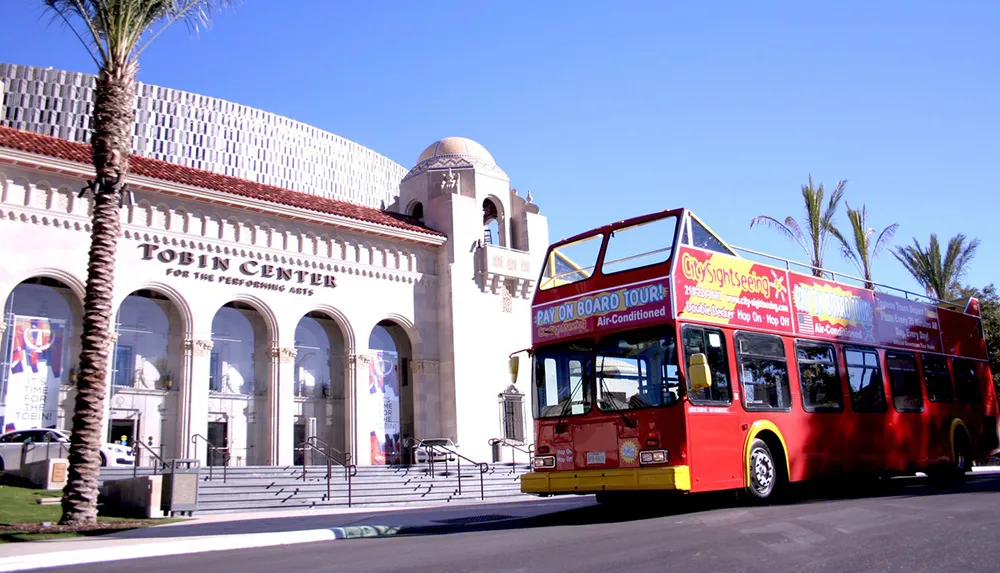 A red double-decker sightseeing tour bus passes in front of the Tobin Center for the Performing Arts under a clear blue sky