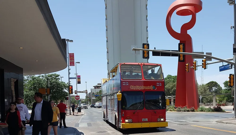 A red double-decker sightseeing bus is stopped at an intersection with pedestrians nearby and a distinctive large red sculpture in the background