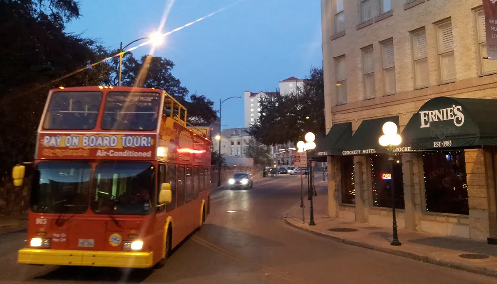 A double-decker tour bus with the sign Pay On Board Tour drives through a street lined with buildings and street lamps as evening sets in