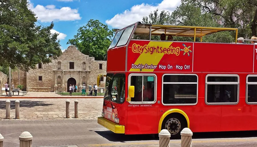 A red double-decker sightseeing bus is parked in front of the historic Alamo mission under a clear blue sky.
