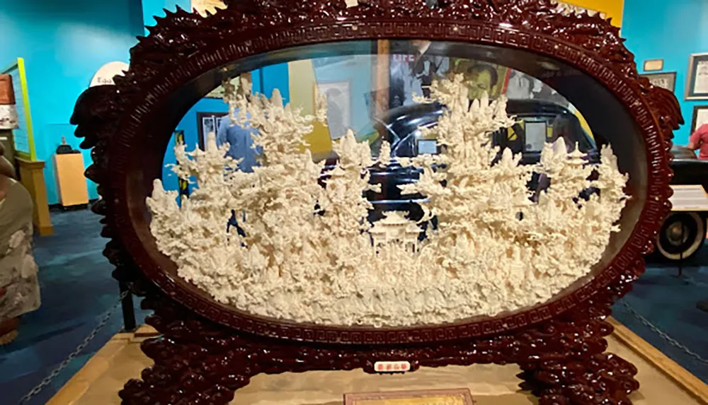 The image displays an intricately carved decorative object resembling a framed oval mirror showcasing a three-dimensional relief of what appears to be a Chinese landscape scene set against the backdrop of a colorful museum exhibit