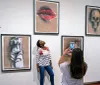 A person admires artwork in a gallery while another takes a photo with their smartphone
