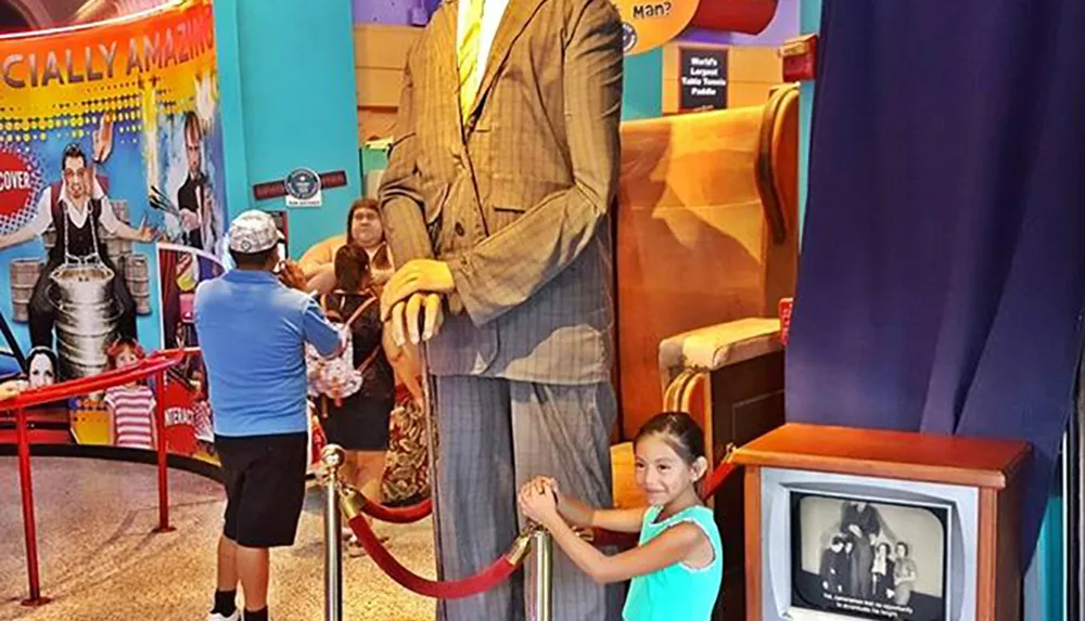 A smiling young girl is standing next to a remarkably tall statue of a man in a suit which dwarfs her in comparison inside what appears to be a museum exhibit celebrating extraordinary human traits or records