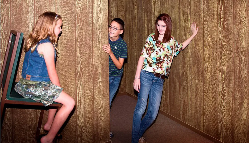 Two children and a teenager appear to interact with an exhibit at an amusement or science center creating an illusion where the teenager looks like a giant due to forced perspective