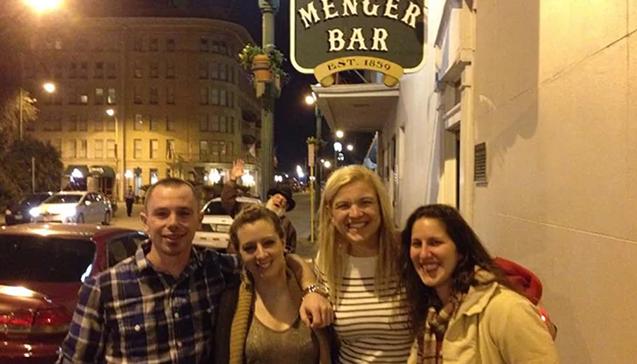 Four people are posing for a photo while smiling on a city street at night in front of the Menger Bar with someone photo-bombing in the background.