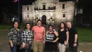 A group of people is posing for a photo at night in front of the historic Alamo mission in San Antonio, Texas, with one individual holding a 