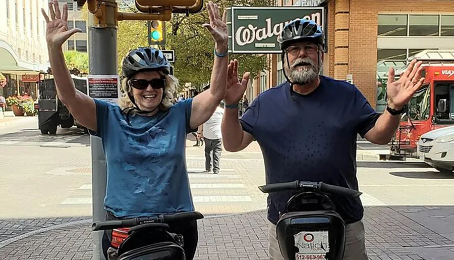 Two people wearing bicycle helmets are joyfully raising their hands in a greeting or celebratory gesture on a city street.