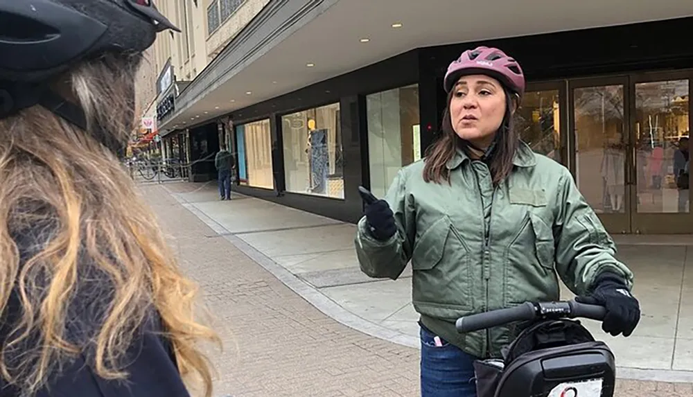 A person in a pink helmet gestures while talking to another person with a bicycle helmet on a city street