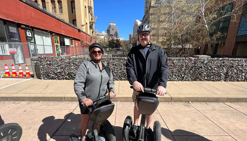 Two people are smiling for the camera while standing on Segways in an urban setting with a wall covered in what appears to be padlocks in the background